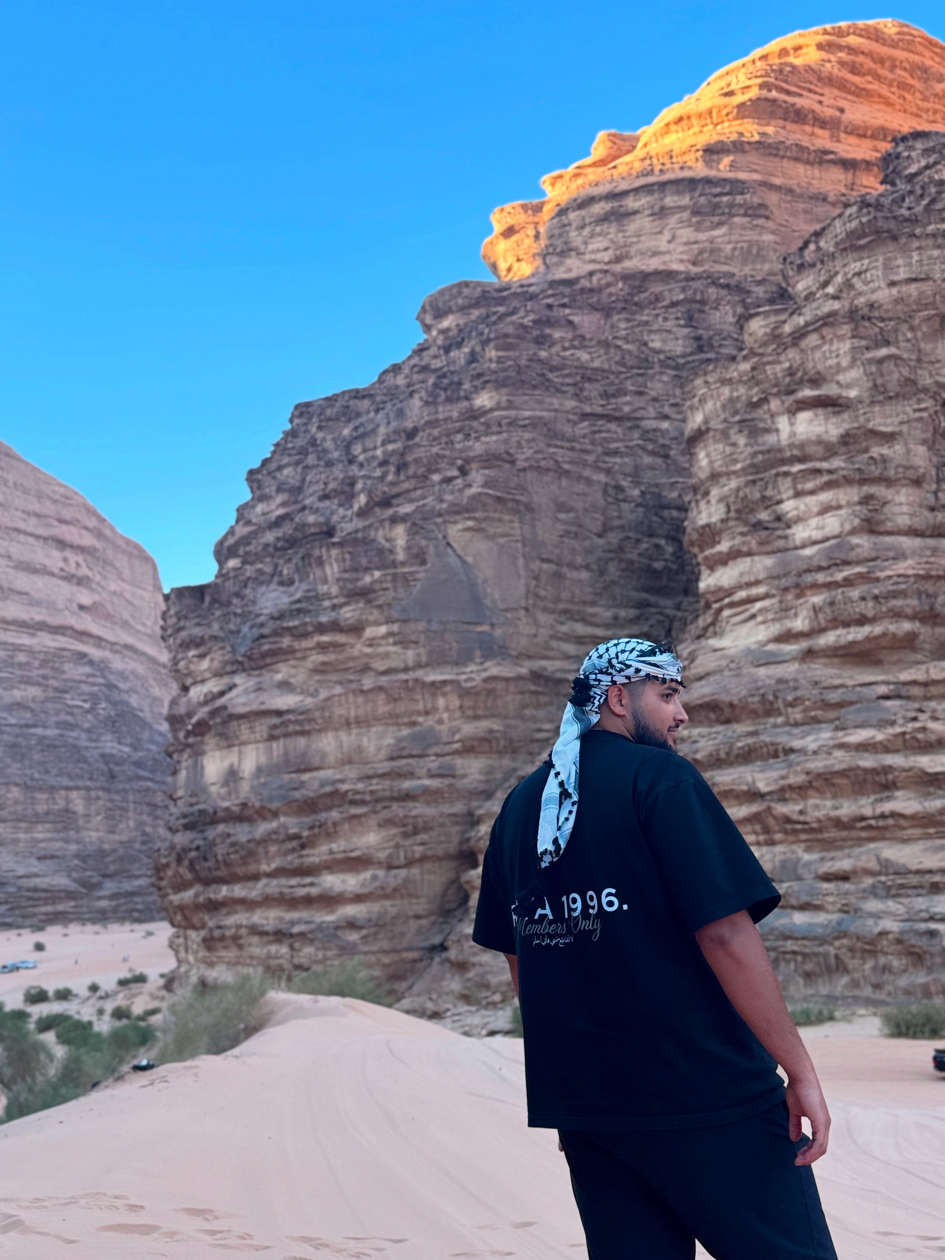 Person standing in front of large rock formations under a clear blue sky. Wearing a black Members Only Tee by uya 1996.