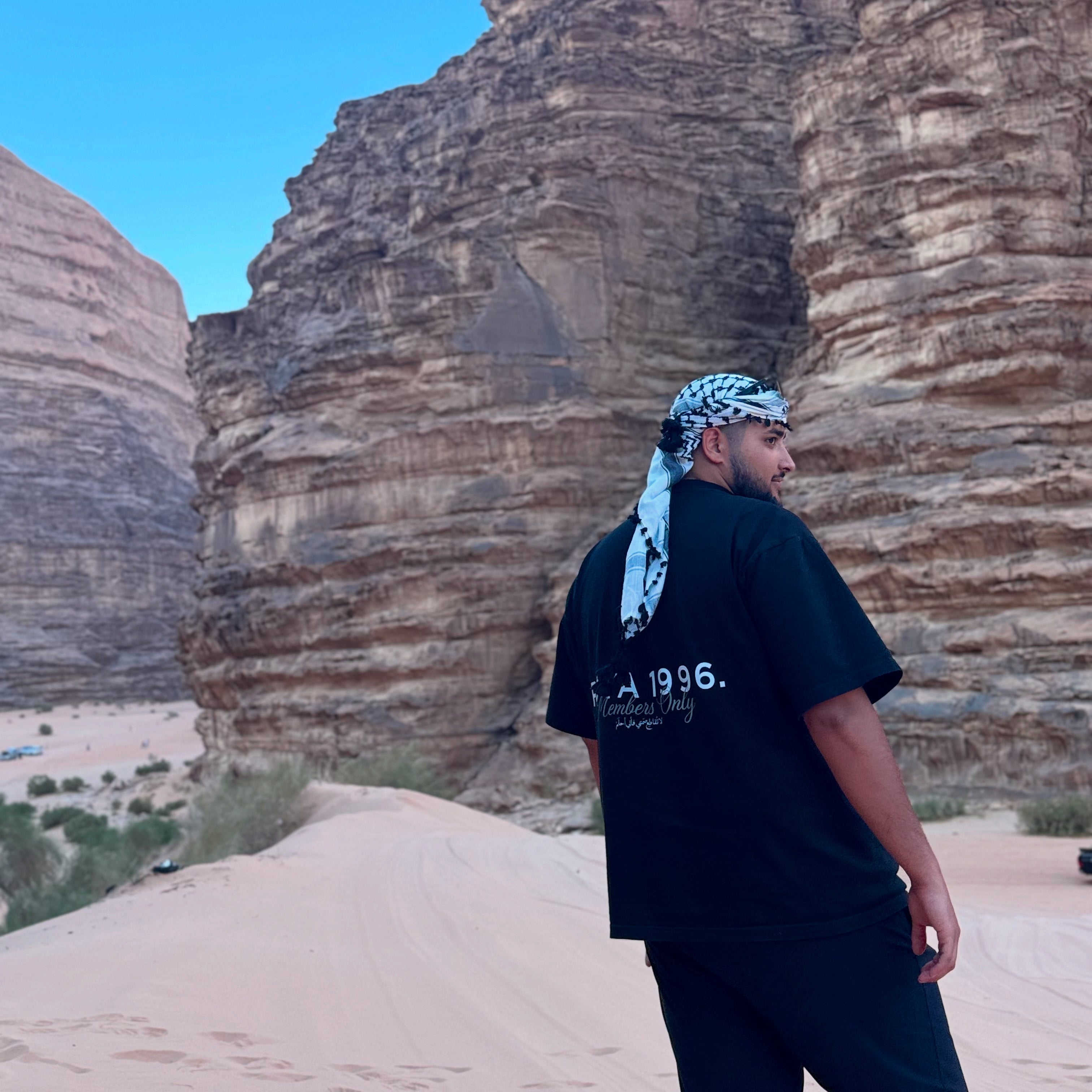 Person standing in front of large rock formations under a clear blue sky. Wearing a black Members Only Tee by uya 1996.