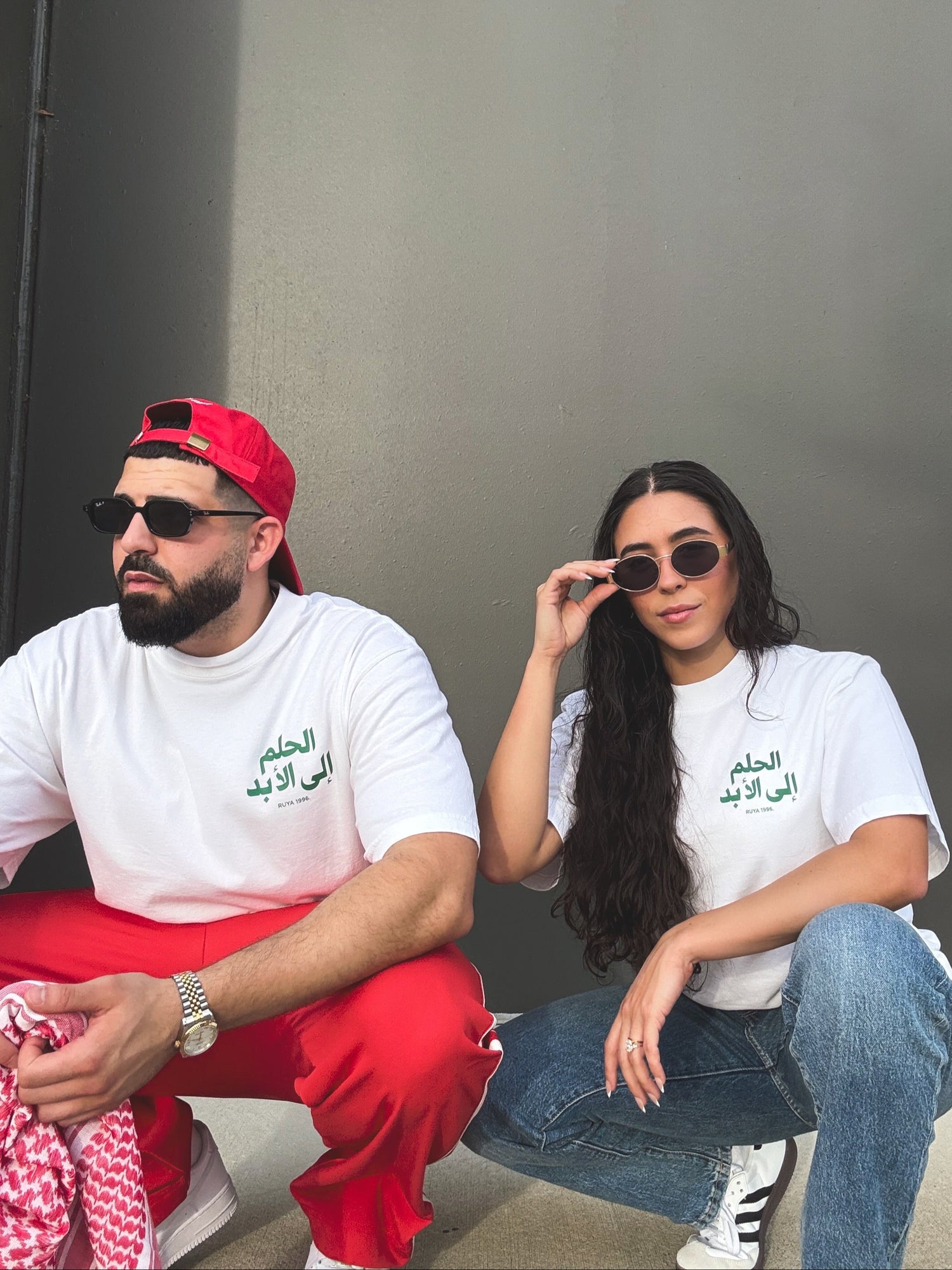 Two people wearing matching white t-shirts with Arabic text, sitting against a gray wall.