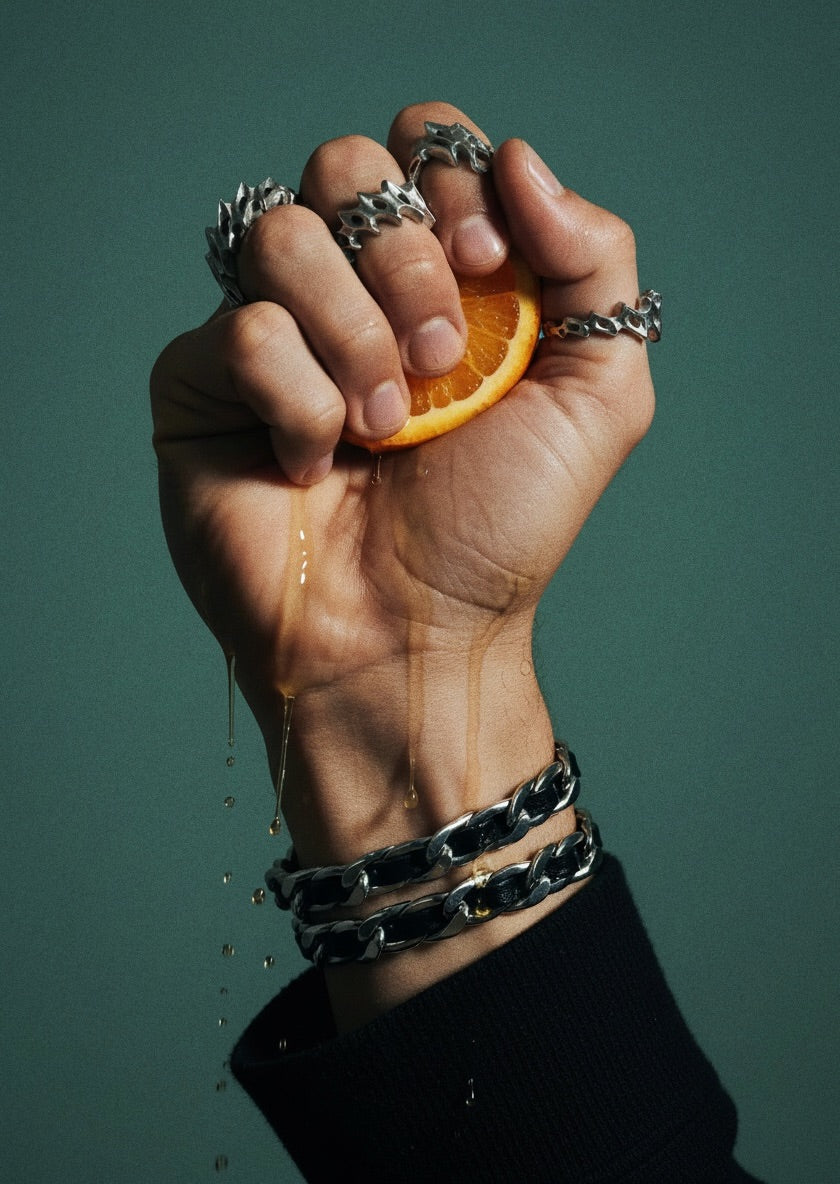 Hand with multiple  unreleased rings and bracelets squeezing an orange against a dark green background